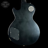 The back of a black Gibson Les Paul Custom electric guitar, featuring a circular chrome 'Gibson Les Paul Custom' emblem on the upper right and a matching chrome cover on the lower left, set against a solid black background.