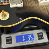 A close-up of a black electric guitar body with gold trim resting on a digital scale displaying 7.35 lbs.