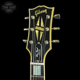 Close-up of the headstock of a Gibson Lvr Rock Custom electric guitar against a black background, featuring a black headstock with gold trim, a gold and black geometric logo, and silver tuning pegs.