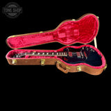 An electric guitar with a black body and red headstock, displayed inside an open brown hard case with bright red velvet lining, set against a solid black background.