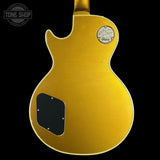 The back of a vintage Gibson Les Paul Custom electric guitar in a metallic gold finish, featuring a black pickguard and a circular Gibson logo badge on the upper back, shown against a solid black background.