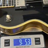 Close-up of a black electric guitar body resting on a digital scale displaying 9.53 oz, with the guitar showing a gold-trimmed edge and a white volume knob.