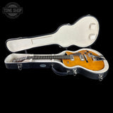 A double-cutaway electric guitar with a natural sunburst finish resting inside an open black hard case with a cream interior, shown against a solid black background. A Tone Shop watermark is visible in the top-left corner.