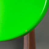 Close-up of a bright lime green curved surface, likely part of a chair or stool, with a small orange stain near the edge and a brown wooden leg visible in the background.