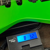 A close-up of a bright green electric guitar body resting on a black Smart Weigh electronic scale, with the scale's blue-backlit display showing a weight reading.