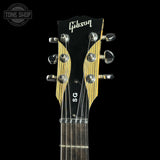 Close-up of a Gibson SG acoustic guitar headstock and upper neck against a solid black background, featuring a black headstock with gold-toned wood grain sides, silver tuning pegs, and a dark fretboard.