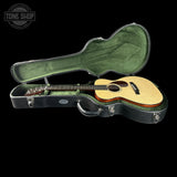 An acoustic guitar with a natural wood body and dark neck, displayed inside an open black hard case with a green velvet interior, set against a solid black background.