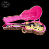 An electric guitar with a cream body and dark headstock, displayed inside an open hard case with a bright pink velvet interior and brown wood-grain exterior, set against a solid black background.