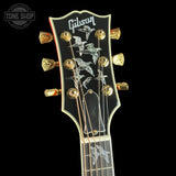Close-up of a Gibson electric guitar headstock featuring a black faceplate with gold tuning pegs, silver bird inlays, and the Gibson logo at the top, set against a solid black background.