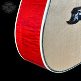 Close-up of an acoustic guitar body showing a bright red flamed maple top edge, a natural beige wood soundboard, and part of a black pickguard on a black background.