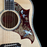 Close-up of an acoustic guitar body showing the natural wood grain, sound hole, strings, and a decorative pickguard with inlaid birds and reeds on a black background.