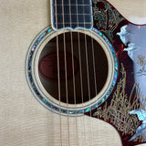 Close-up of an acoustic guitar body showing the sound hole, six strings, a light beige wood neck, and a dark reddish-brown pickguard decorated with silver bird illustrations and gold reeds.