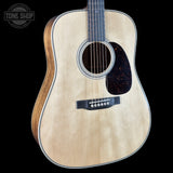 An acoustic guitar with a natural light wood finish and dark brown sides, shown against a solid black background. A Tone Shop watermark is visible in the top-left corner.