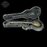 An electric guitar with a sunburst finish resting inside an open hard guitar case on a black background. The case is gray with a black velvet interior.