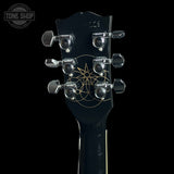 Close-up of a guitar headstock with six silver tuning pegs and a gold geometric star-and-circle logo on a black background. A Tone Shop watermark is visible in the upper left corner.