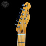 Close-up of a Fender Telecaster electric guitar headstock in a natural maple finish, showing six black tuning pegs and the Fender Telecaster logo, set against a solid black background.