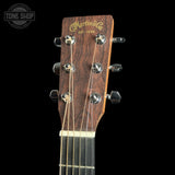 Close-up of the headstock of a Martin & Co acoustic guitar, showing the dark wood grain, gold script logo, and six silver tuning pegs against a black background.