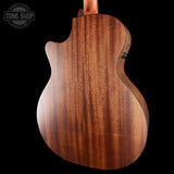Close-up of the back of an acoustic guitar body made from rich, warm brown wood with prominent grain patterns, set against a solid black background. A Tone Shop watermark is visible in the top-left corner.