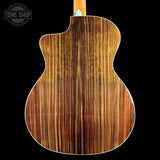 Close-up of the back of an acoustic guitar showing a rich wood grain finish in warm brown and amber tones, set against a solid black background with a Tone Shop watermark in the top left corner.