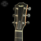 Close-up of a Taylor acoustic guitar headstock with six tuning pegs, strings, and a dark wood finish against a black background.