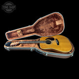 An acoustic guitar with a natural wood finish sits inside an open hard guitar case with a brown velvet interior, shown against a solid black background. A Tone Shop watermark is visible in the top-left corner.