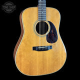 An acoustic guitar with a natural wood finish and a dark tortoiseshell pickguard, shown against a solid black background with a Tone Shop watermark in the top left corner.