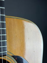 Close-up of an acoustic guitar body showing the natural wood grain, sound hole rosette, and part of the dark fretboard against a dark background.