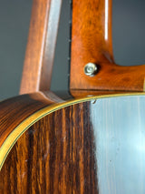 Close-up detail of an acoustic guitar's wooden body, showing the rich dark brown wood grain, a gold-toned binding edge, and part of the neck with a metal tuning peg visible against a dark gray background.