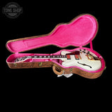A white electric guitar with gold hardware displayed inside an open hard case with a bright pink velvet interior and brown cork-look trim, photographed on a black background. A Tone Shop watermark appears in the upper left corner.