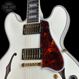 Close-up of a white electric guitar body with a red and black tortoiseshell-style pickguard, black humbucker pickups, gold hardware, and a dark fretboard, shown against a black background. A Tone Shop watermark is visible in the upper left.