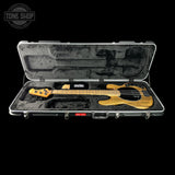 A natural wood five-string bass guitar displayed inside an open silver hard case with black foam lining, set against a solid black background. A Tone Shop watermark is visible in the top-left corner.