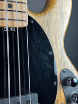 Close-up of a bass guitar body showing a natural wood finish, black pickguard with visible scratches, control knobs, and part of the fretboard.