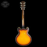 A vintage-style electric guitar shown from the back, featuring a classic sunburst finish that transitions from bright yellow-orange in the center to dark brown at the edges. The headstock is dark brown wood with six silver metal tuning pegs.