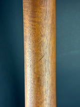 A close-up of a cylindrical wooden pole or post showing a warm medium-brown wood grain texture against a dark blue background.