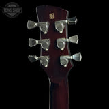 Close-up of the headstock of a dark reddish-brown acoustic guitar against a black background, showing six silver tuning pegs and a small gold logo badge. A Tone Shop watermark is visible in the top left corner.