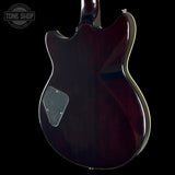 Close-up of the rear body of an electric guitar against a black background, showing a glossy dark reddish-brown finish, a silver hardware plate, and a thin gold edge trim. A Tone Shop watermark is visible in the top-left corner.