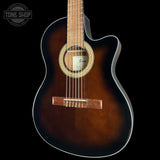 A close-up of a cutaway acoustic guitar against a solid black background, showcasing its dark brown sunburst finish, natural wood neck, and gold-toned rosette.