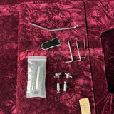 A set of metal hardware tools and small parts laid out on a deep burgundy velvet surface, including a long curved metal rod, several L-shaped wrenches, a small plastic bag with a metal tube, two small metal connectors, and a tan leather tag.