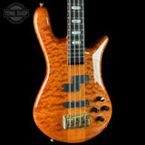 A five-string electric bass guitar with a glossy orange flamed maple top, black hardware, and a black neck, shown against a solid black background with a Tone Shop watermark in the upper left corner.