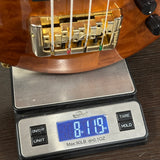 Close-up of a wooden bass guitar body resting on a digital scale showing a weight of 8.19 lbs, with the bridge and strings visible.