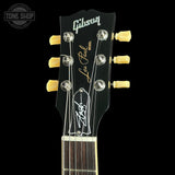 Close-up of a Gibson Les Paul Model electric guitar headstock, featuring a black headstock with gold and white lettering, six gold tuning pegs, and a white Gibson logo, shown against a solid black background.