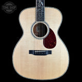 A front-facing acoustic guitar with a natural wood finish and dark pickguard, shown against a solid black background with a Tone Shop logo in the top-left corner.