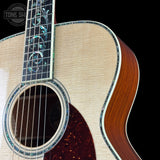 Close-up of an acoustic guitar body and neck, showing a natural wood grain top, dark brown sides, decorative inlay around the soundhole and fretboard, and a black background. A Tone Shaper logo is visible in the upper left corner.