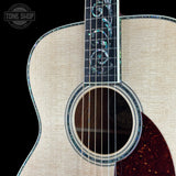Close-up of an acoustic guitar body and neck with a light natural wood grain top, dark fretboard, iridescent inlay, and a dark reddish-brown pickguard on a black background.
