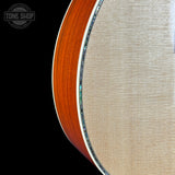 Close-up detail of an acoustic guitar's side, showing the warm orange-brown wood body edge, a silver inlay, and a light beige fabric-covered fingerboard against a black background. A Tone Shop watermark is visible in the top-left corner.