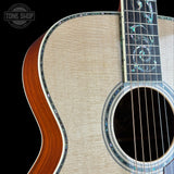 Close-up of an acoustic guitar body and neck against a black background, showing a natural wood grain top, decorative inlay around the soundhole and edge, and a dark fretboard with inlaid fret markers. A Tone Shop watermark appears in the upper left.