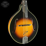 A close-up of a mandolin against a solid black background, showcasing its sunburst finish, gold-toned binding, and silver metal bridge. A Tone Shop watermark is visible in the top-left corner.