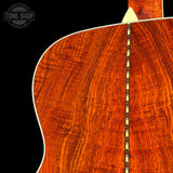 Close-up of the back of an acoustic guitar showing a rich, flamed wood grain finish in warm reddish-brown tones, with a decorative inlay running vertically down the center. A Tone Shop watermark is visible in the top-left corner against a black background.