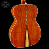 Close-up of the back of an acoustic guitar showing a rich, warm reddish-brown wood grain finish with a light natural wood center stripe and white binding, set against a solid black background. A Tone Shop watermark is visible in the top left corner.