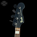 Close-up of a Fender bass guitar headstock in black with silver tuning pegs and a gold Fender logo, shown against a black background with a Tone Shop watermark in the upper left.
